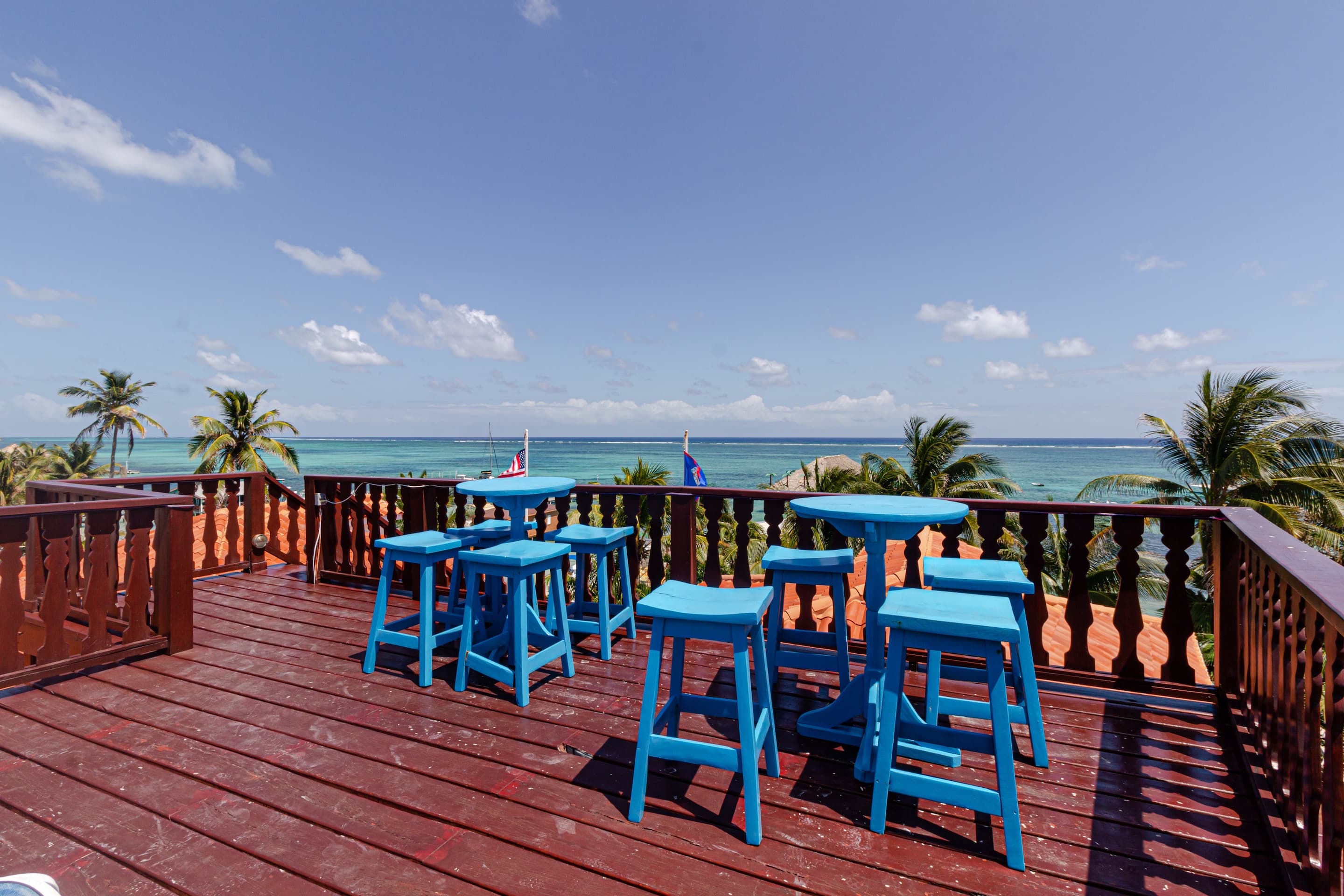 A wooden rooftop deck with blue bar stools and small tables overlooking the ocean and palm trees und
