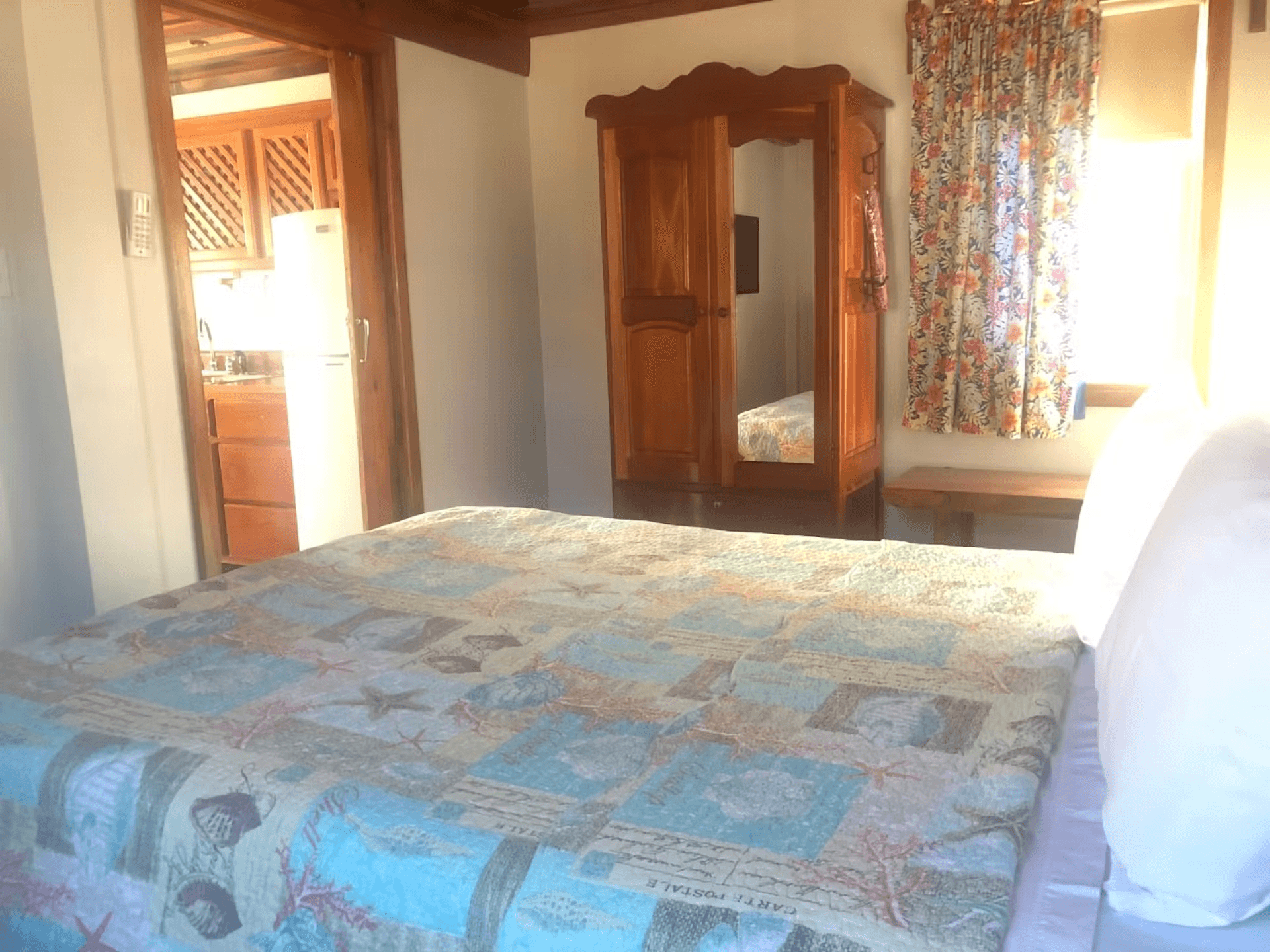 Bedroom with queen bed, wooden armoire, doorway leading into kitchen, and sunlight streaming through