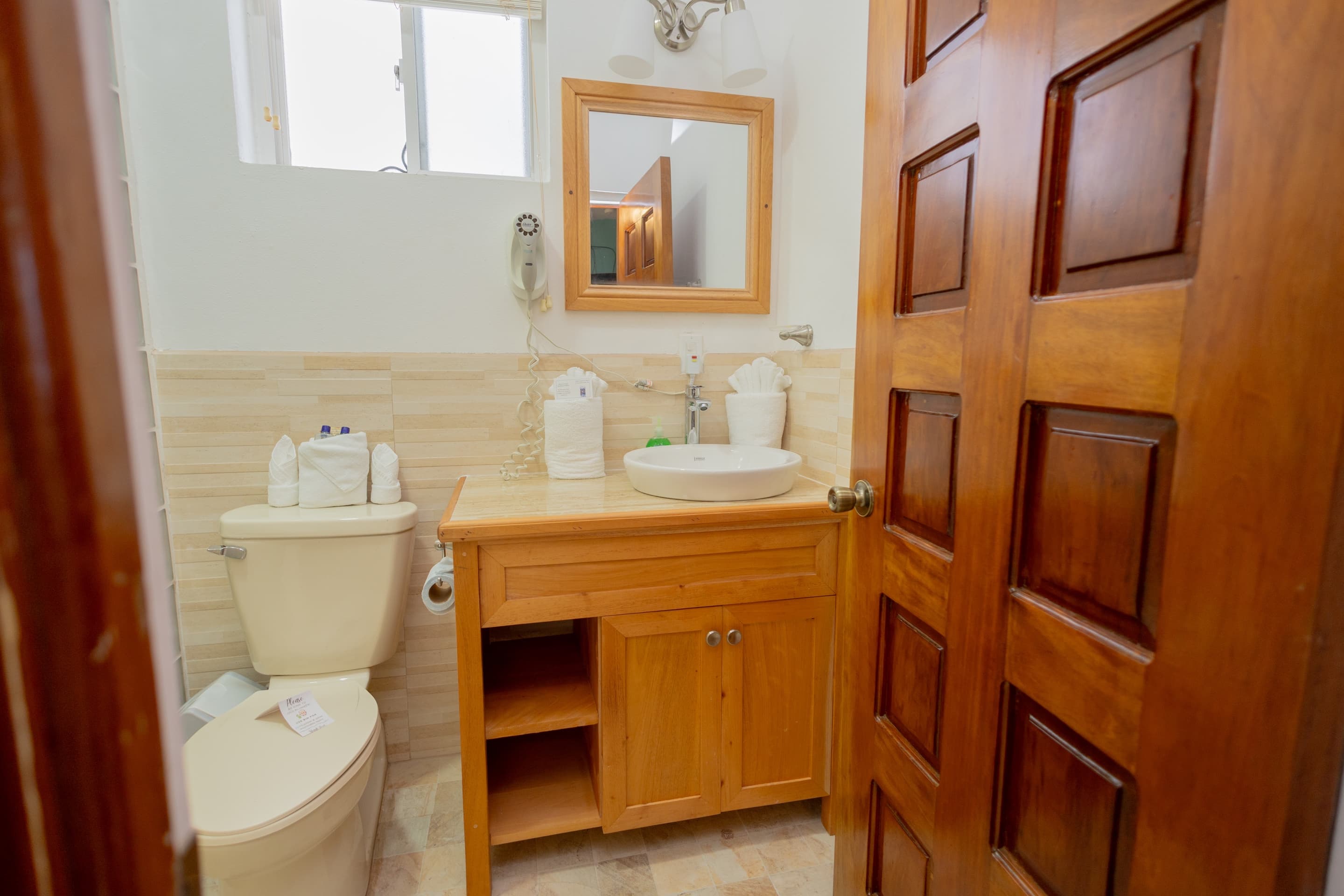 A compact bathroom with light tiled walls, a wooden-framed mirror above a sink with a wooden cabinet