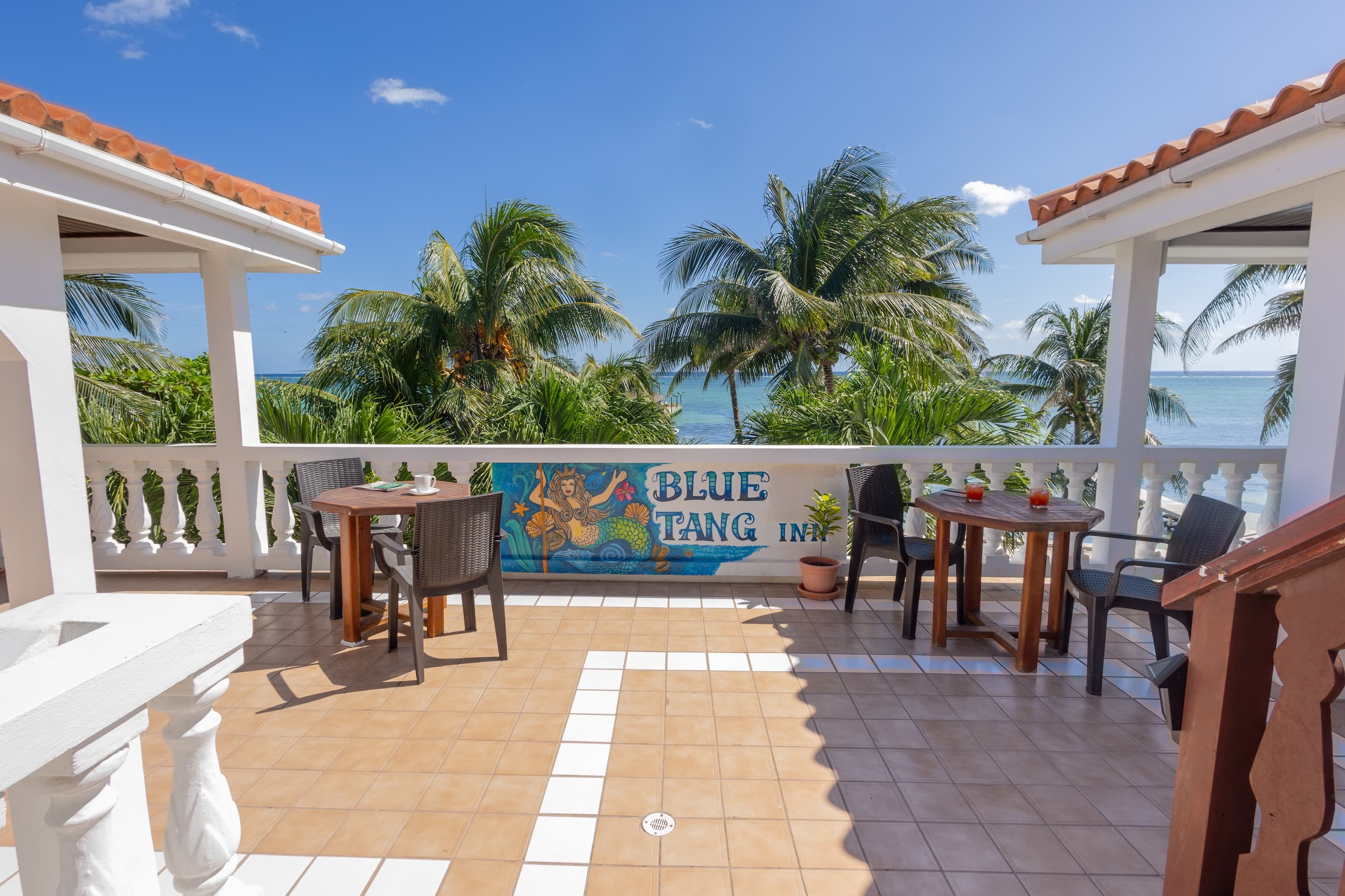A rooftop patio with tables and chairs, white railings, and a view of the ocean and palm trees under