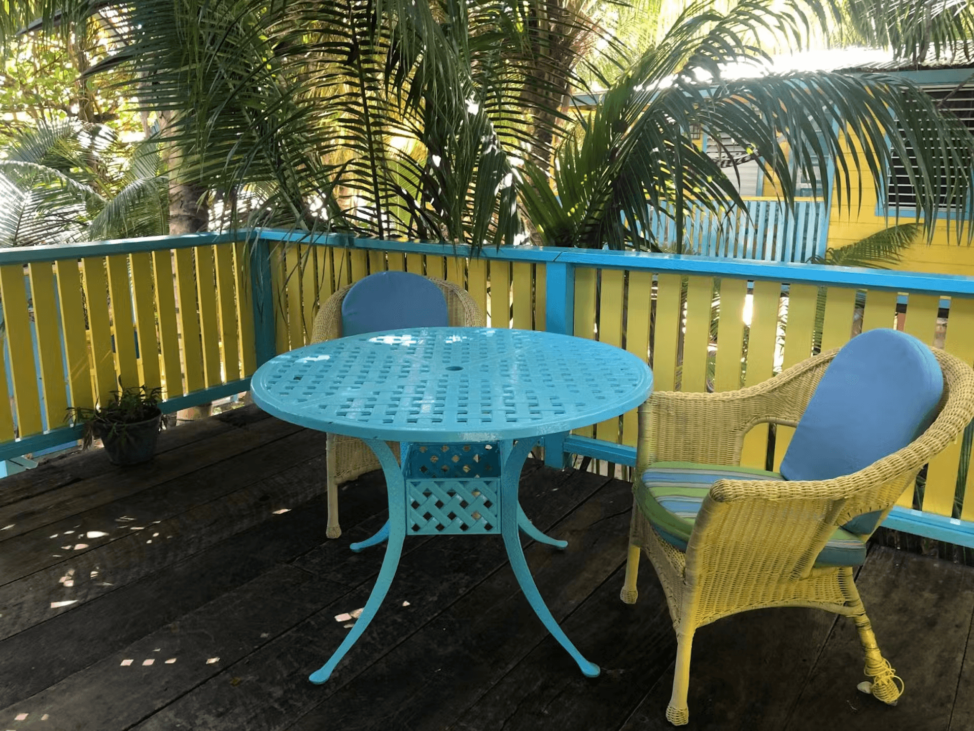 A balcony with yellow and blue rails and two yellow chairs with a round bright blue table, palm tree