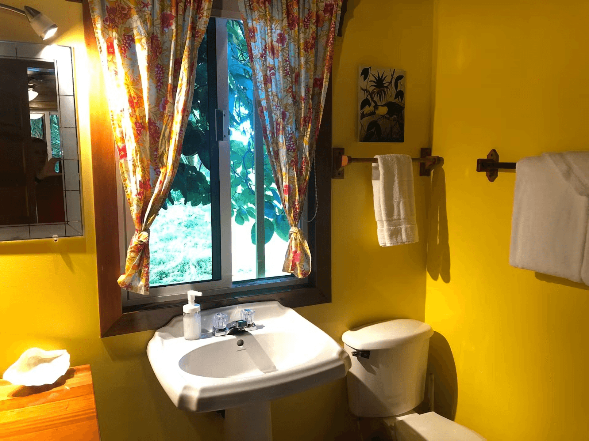 Bathroom painted bright yellow, a white pedestal sink with curtained window above, toilet, and white
