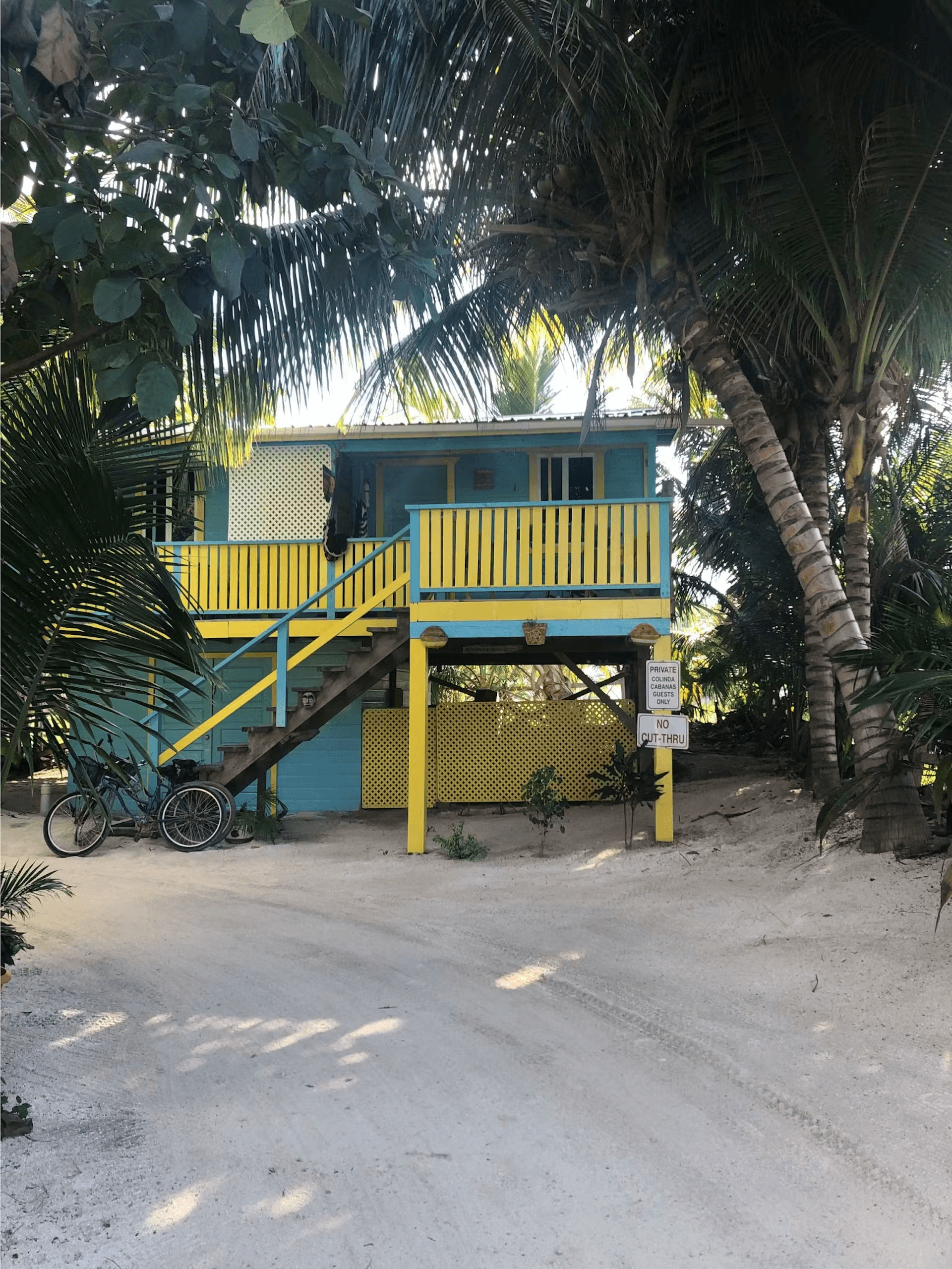 Exterior of brightly painted blue and yellow cabana with stairs leading to balcony, surrounded by sa