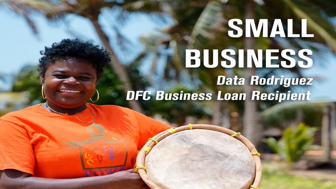 A smiling woman in an orange shirt holds a drum outdoors, representing a small business loan recipient.