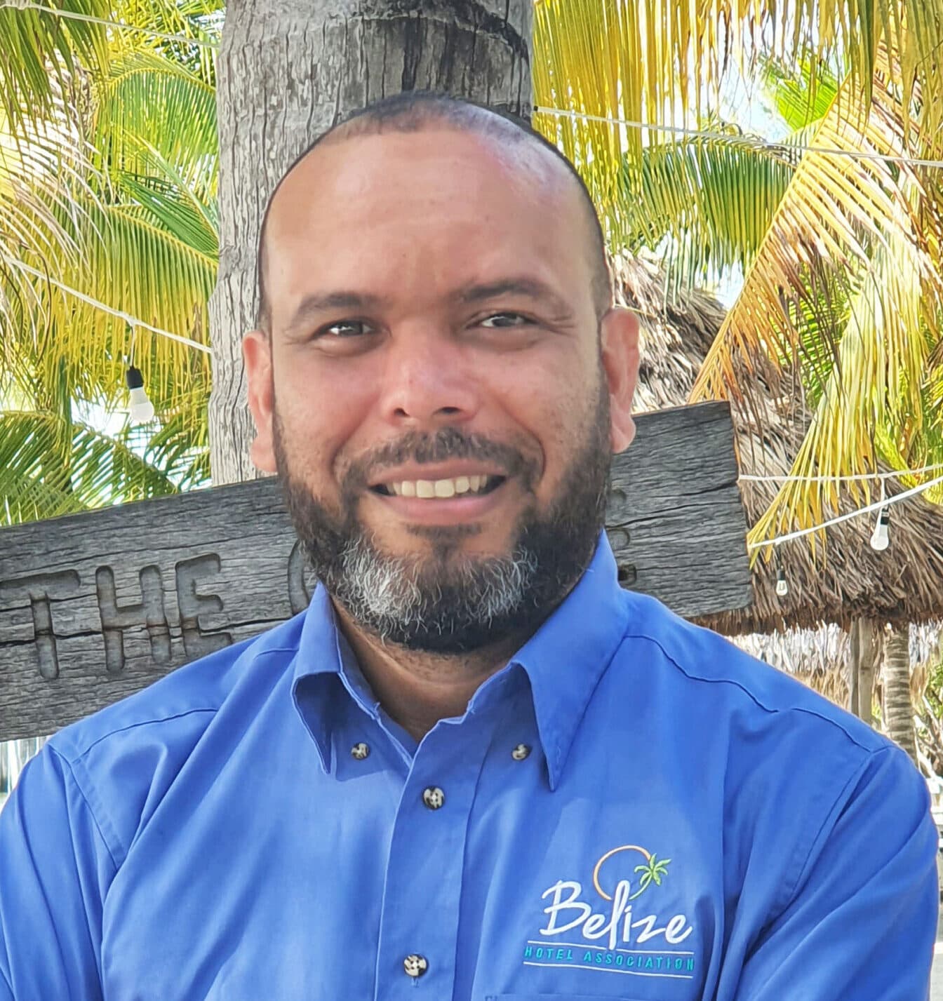 A man with a beard smiles in front of tropical foliage and a wooden sign. A man with a beard smiles in front of tropical foliage and a wooden sign.
