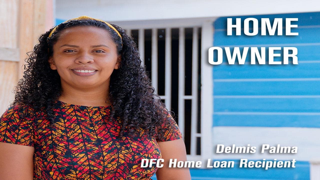 A smiling woman stands in front of a blue house, identified as a DFC home loan recipient.