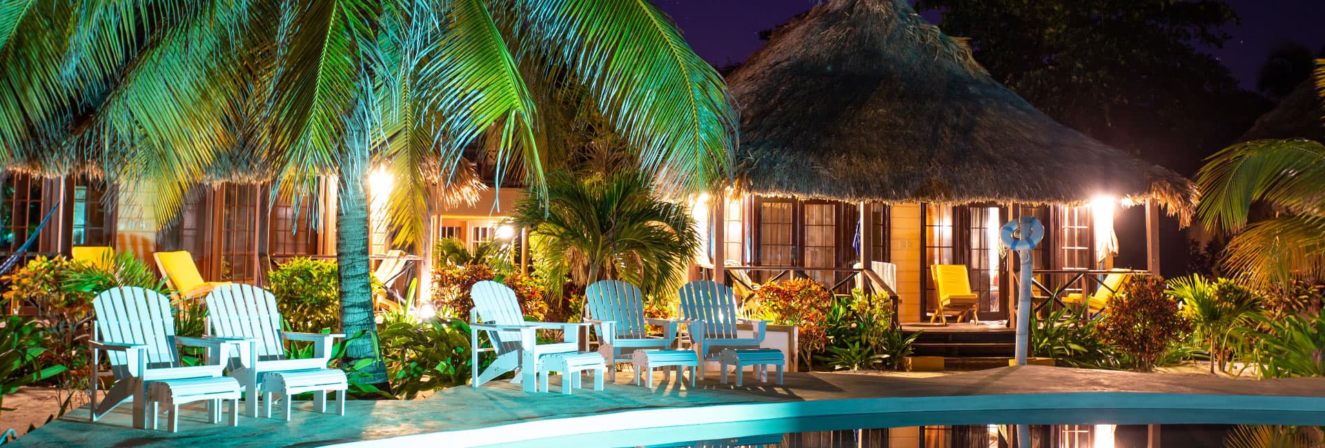 A tranquil resort scene featuring palm trees, a thatched-roof bungalow, and white lounge chairs by a pool at night.