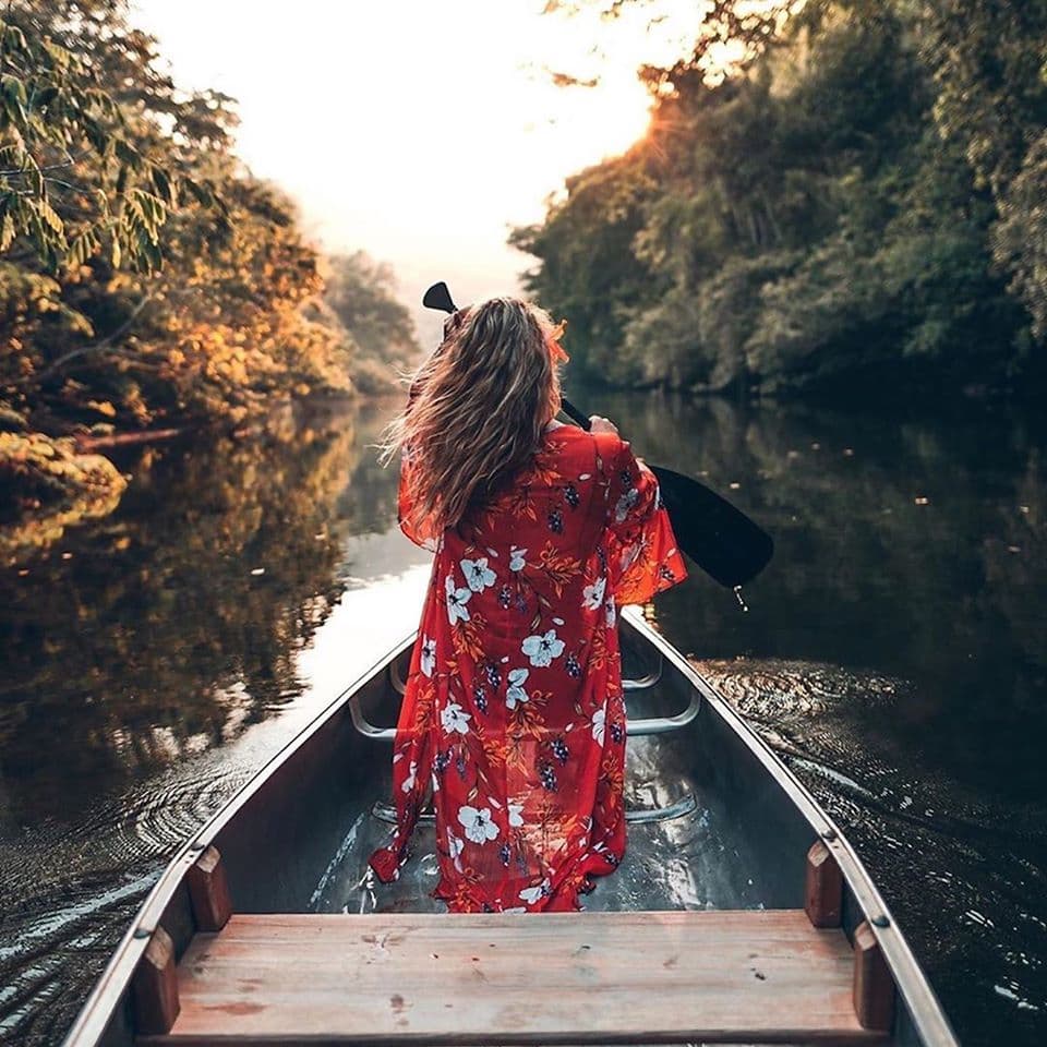 A woman in a floral red robe paddles a canoe along a tranquil river at sunset.