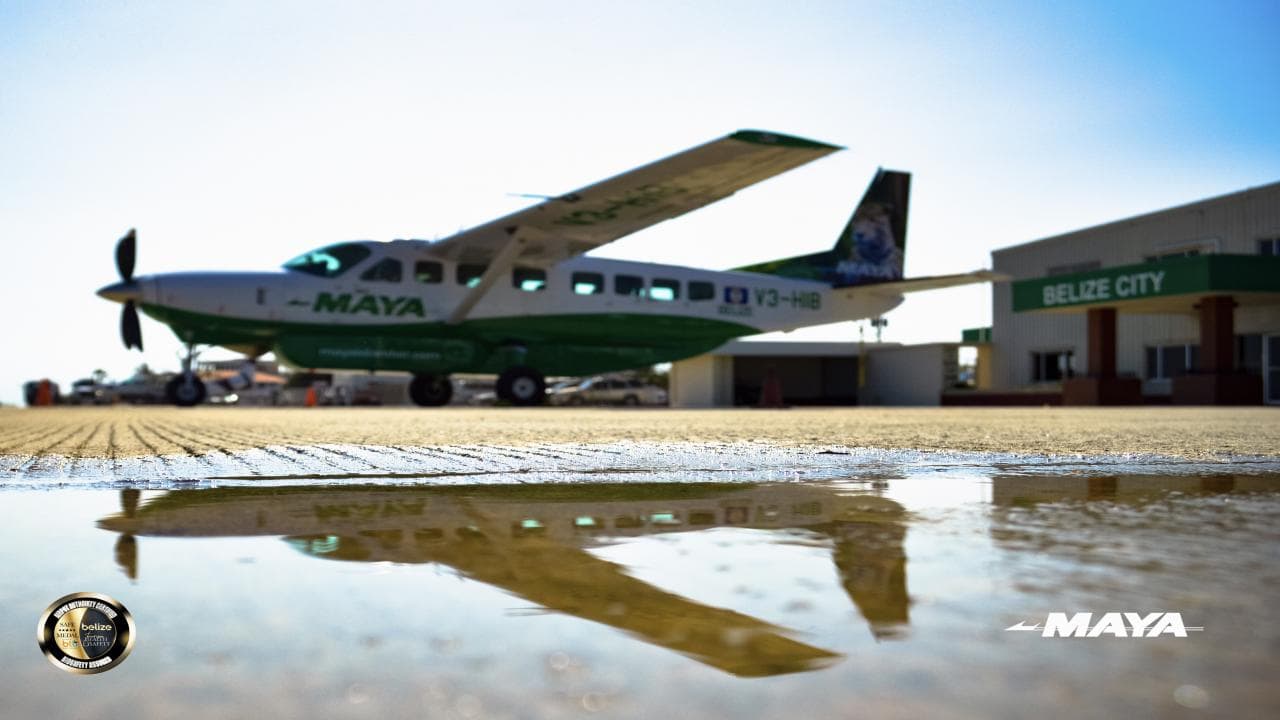 A green and white airplane is parked on the tarmac, reflecting in a puddle, with a terminal building in the background.