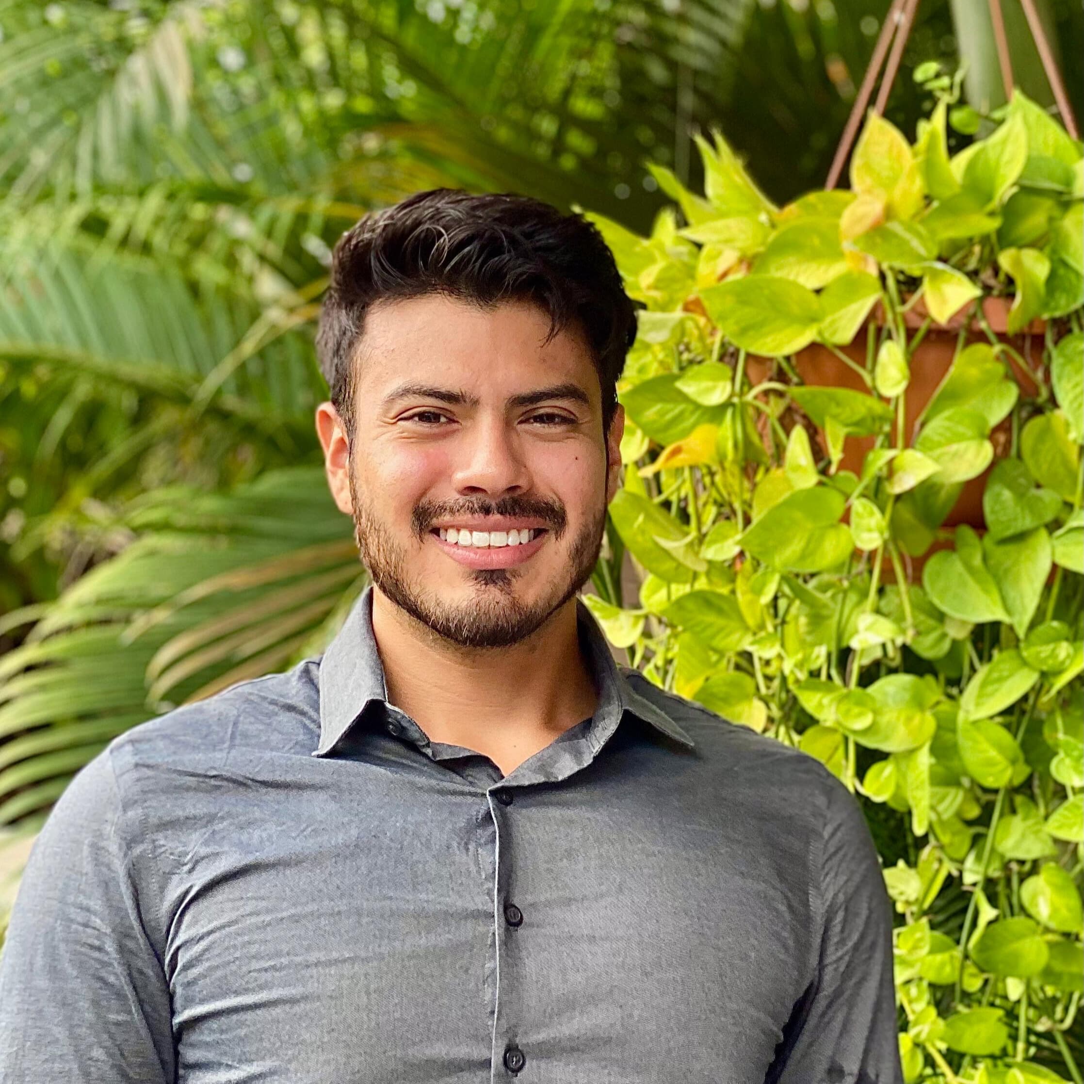 A smiling man in a gray shirt stands in front of lush green plants.