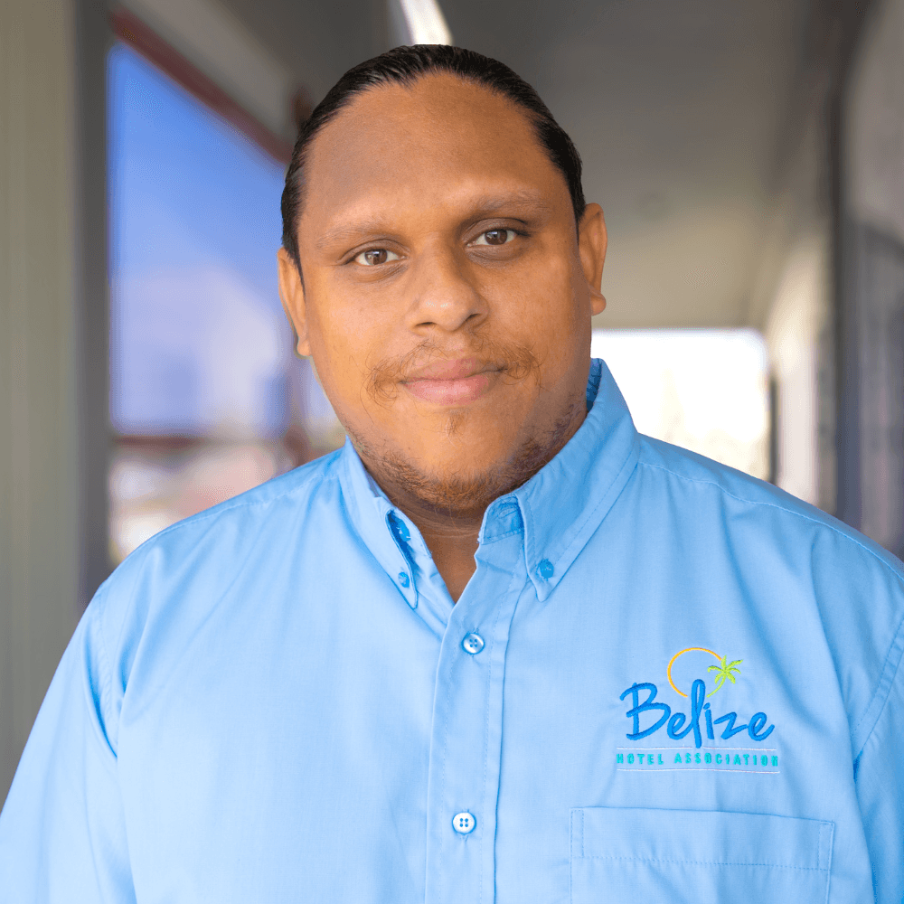 A man in a blue button-up shirt with a "Belize Hotel Association" logo smiles at the camera.
