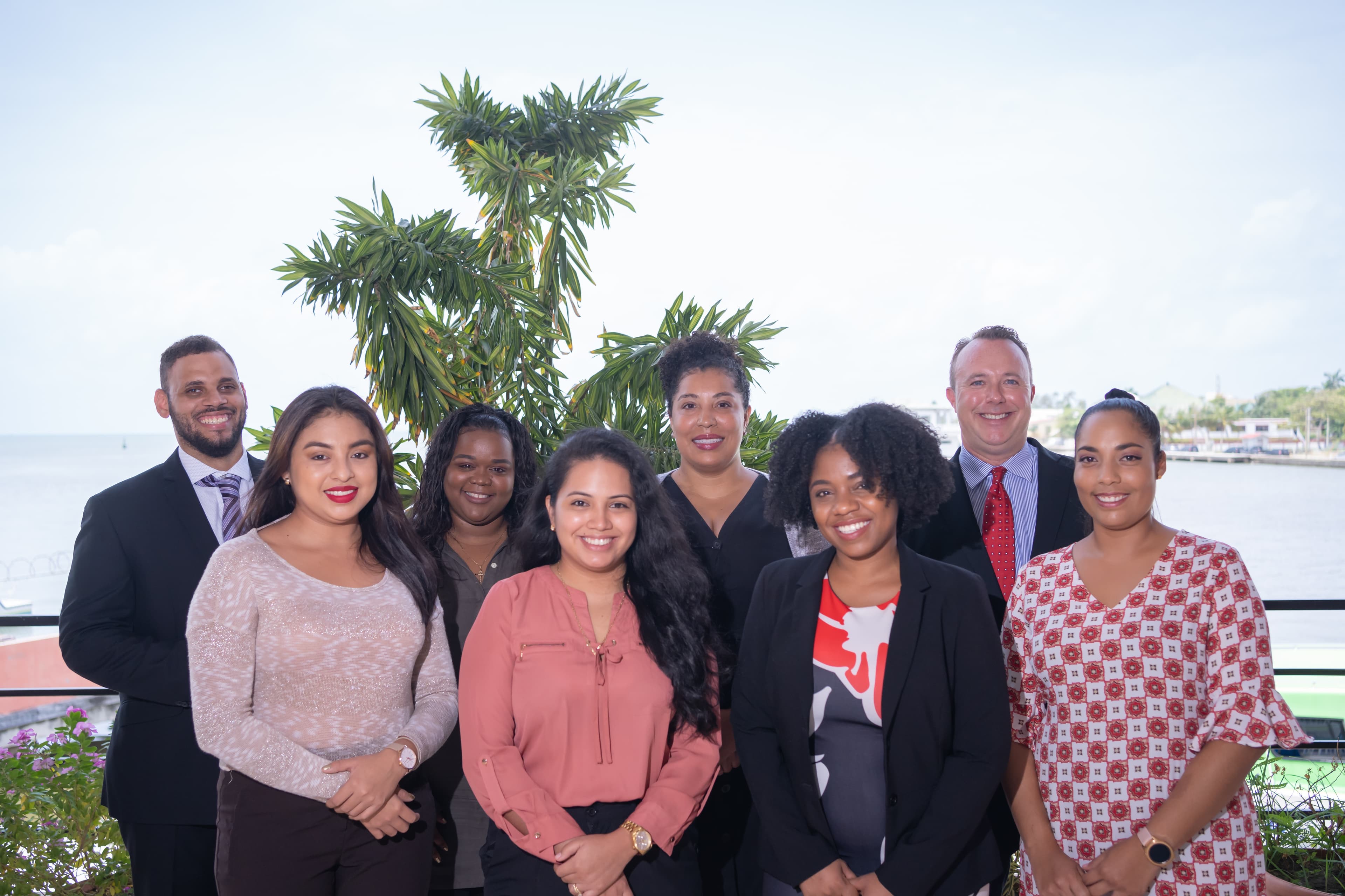 A diverse group of eight professionals poses together outdoors with a waterfront backdrop.