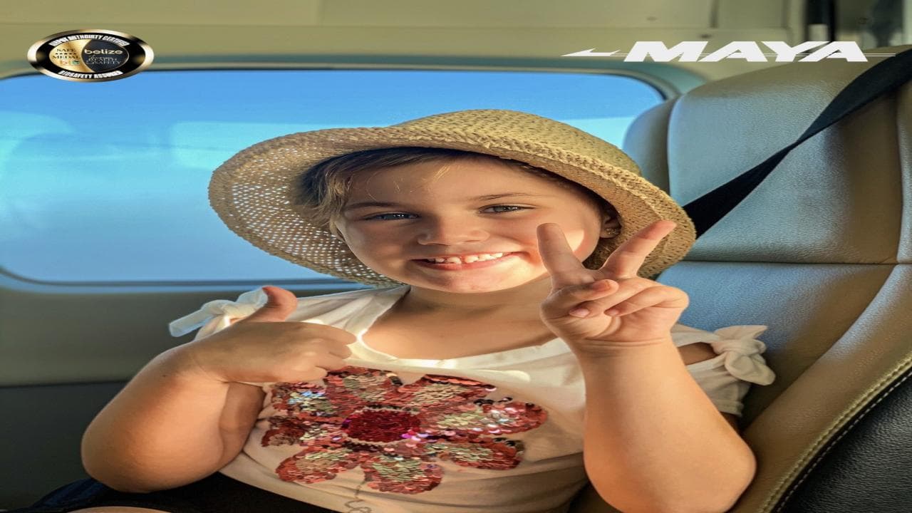 A smiling girl in a straw hat makes a peace sign while sitting by a window.