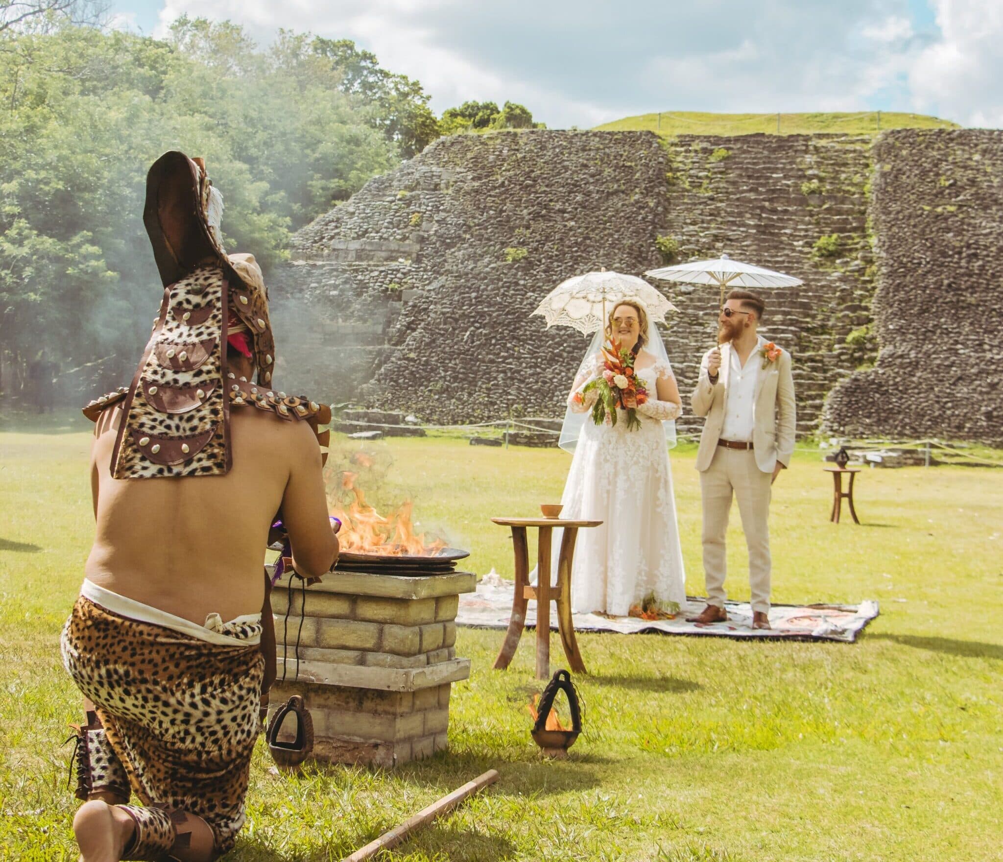 A wedding ceremony taking place outdoors with a couple in formal attire and a person in traditional dress near a fire. A wedding ceremony taking place outdoors with a couple in formal attire and a person in traditional dress near a fire.
